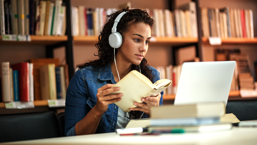 A woman wearing headphones is reading a book and looking at her laptop screen _ Blended Mobility Programmes